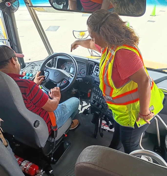 A school bus driver trainer in a high-visibility safety vest provides behind-the-wheel instruction to a driver seated at the controls of a school bus.