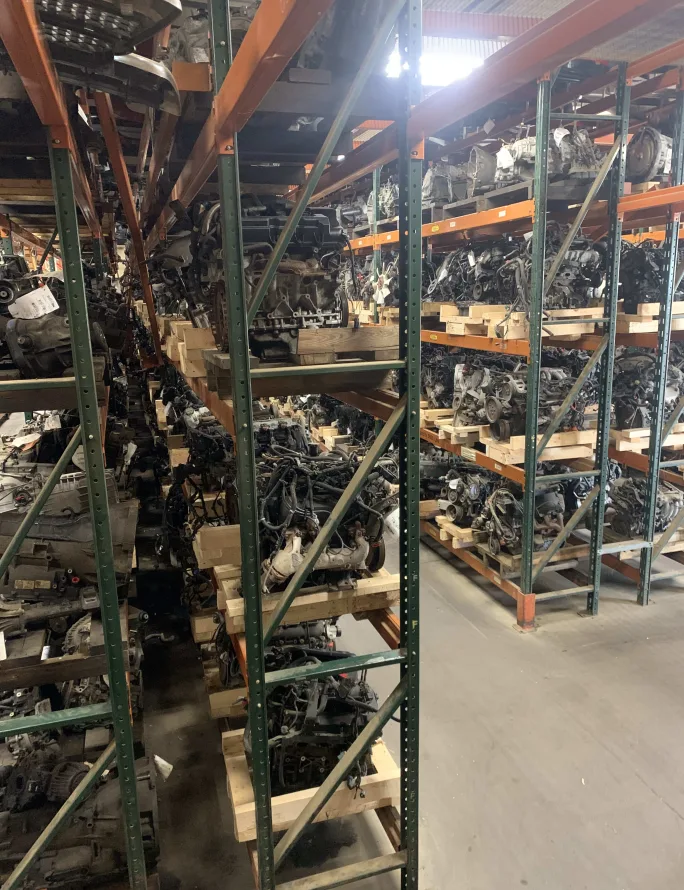 Rows of industrial shelving inside a warehouse are filled with salvaged engines, transmissions, and vehicle components stored on wooden pallets for reuse and recycling.