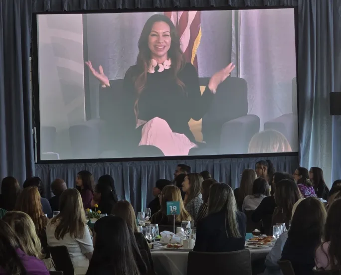 Jennie Abarca on a large video screen in front of a group of people sitting at tables