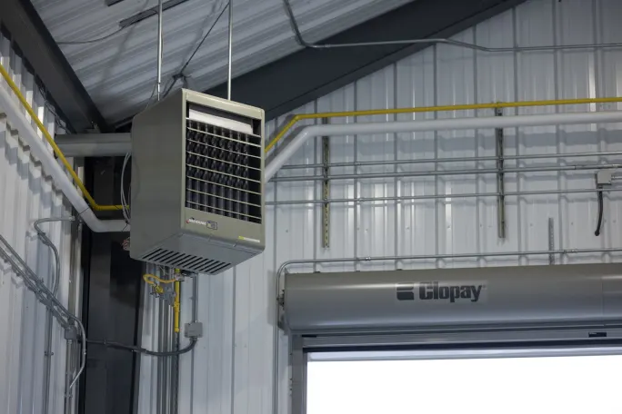 Ceiling-mounted Modine unit heater installed near an overhead garage door, with visible gas piping and ventilation equipment inside a metal-walled maintenance facility.