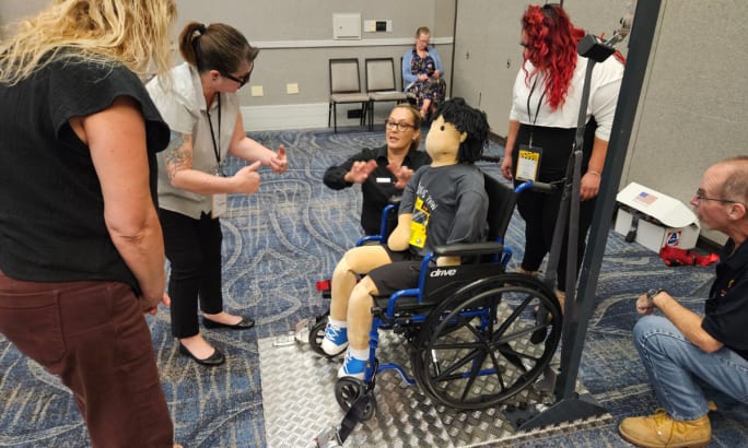 Group training session demonstrating wheelchair securement: an instructor guides participants as they position a wheelchair with a seated mannequin on a platform, showing proper tie-down and safety procedures while others observe.