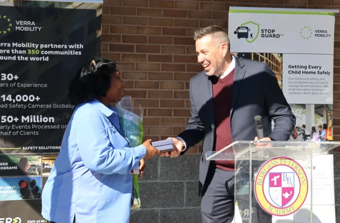A man at a podium presents an award to an Atlanta Public Schools bus driver during a Verra Mobility Stop Guard school bus safety event outside Burgess-Peterson Academy.