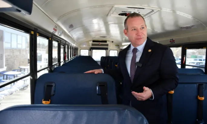 Josh Gottheimer stands inside a school bus and gestures at a seat with lap-and-shoulder belts.