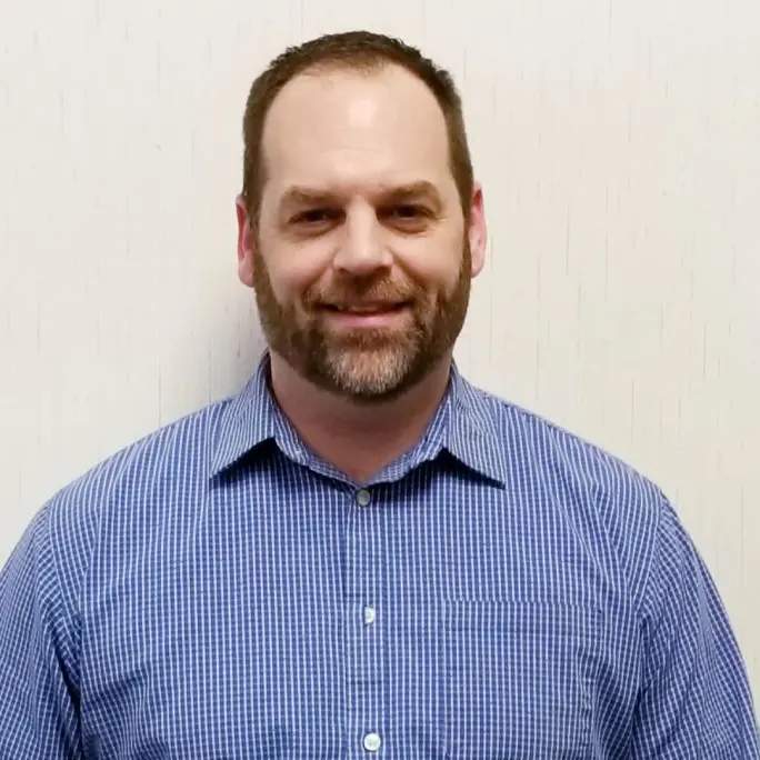 Headshot of Jamie Tuinstra of Modine Manufacturing, smiling in a blue checkered button-down shirt against a light-colored background.