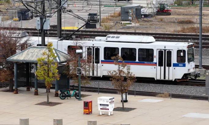 A Denver Regional Transportation District (RTD) light rail train passes a ballot drop-off box near a transit station platform.