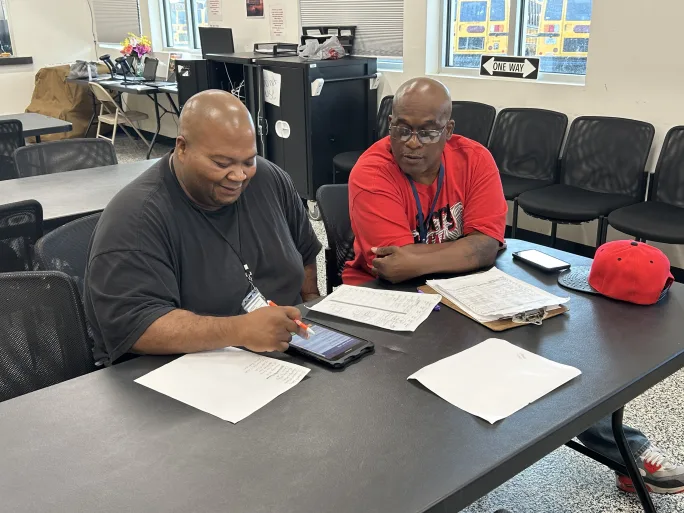 Two men sit at a table with papers in front of them.