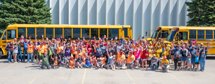 Large group of school transportation employees and families wave at the camera while gathered in front of three yellow school buses.