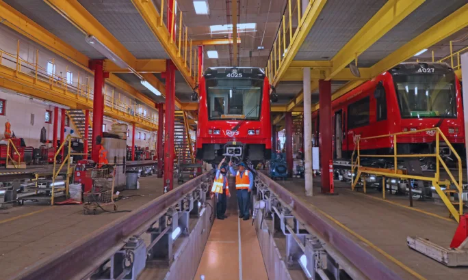 San Diego Metropolitan Transit System employees work beneath a trolley in an MTS rail maintenance facility, with another trolley positioned on an adjacent track.