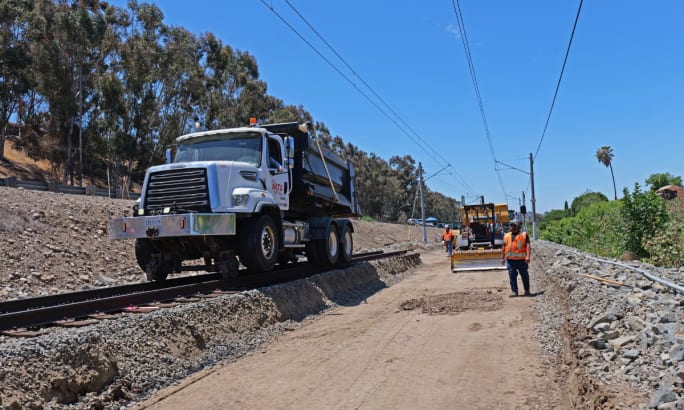 San Diego Metropolitan Transit System crews work alongside construction equipment and a maintenance truck on a trolley rail line.