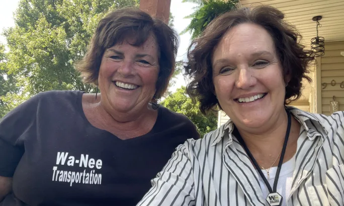 Two Wa-Nee Schools transportation staff members smile for a selfie outdoors, with one wearing a black Wa-Nee Transportation shirt and the other wearing a striped button-down shirt and Wa-Nee badge.