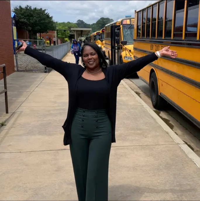 a woman stands next to school buses on a sidewalk with her arms up