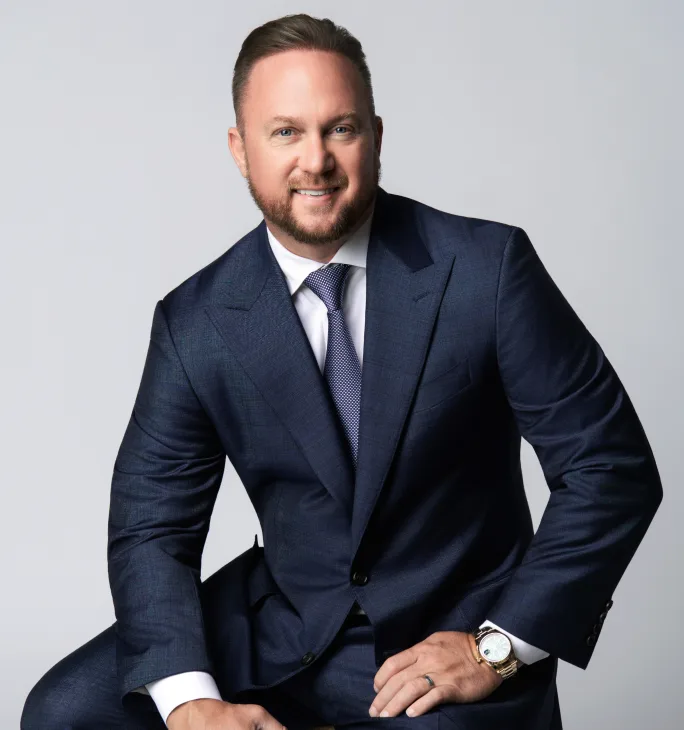 Professional portrait of a smiling, bearded man wearing a navy blue suit, white shirt, and patterned blue tie, seated on a stool against a light gray background.