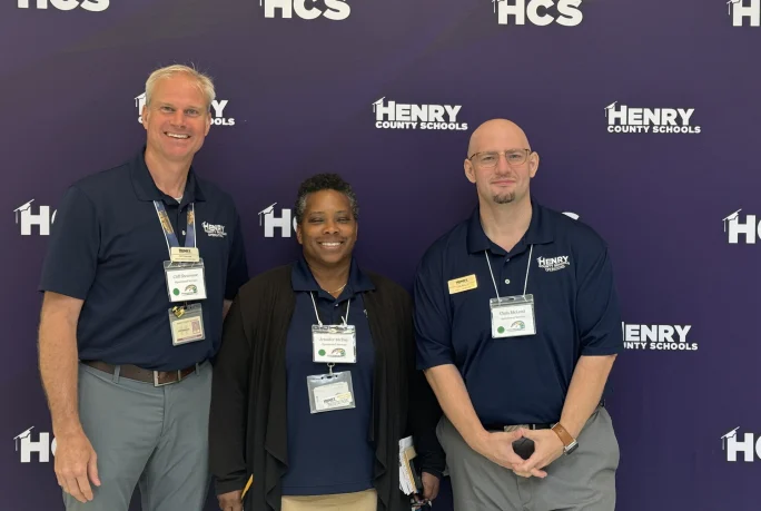 three people from Henry County Schools stand in front of a purple step and repeat banner