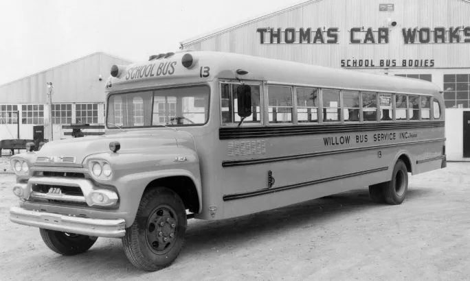 A black and white photo of an early school bus in front of a Thomas Car Works building.