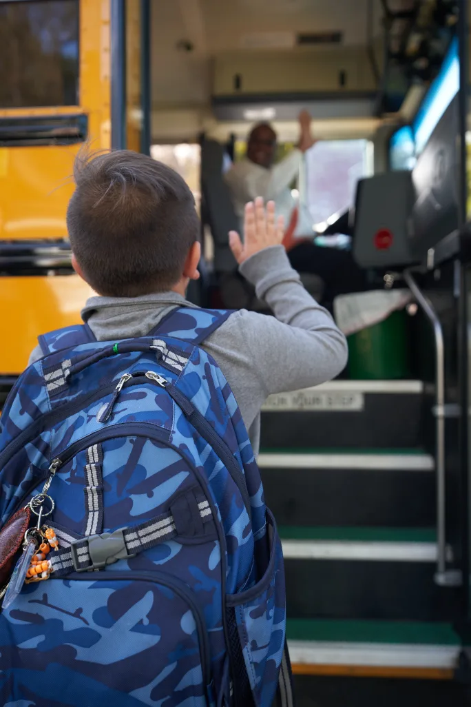 A young student waving at a bus driver through open school bus doors.