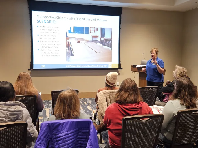 A presenter leads a training session on transporting children with disabilities, speaking to an audience seated in a classroom-style setting while a slide on special education law is displayed.