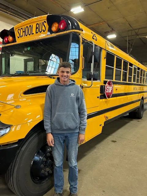a young man stands in front of a school bus inside a maintenance shop