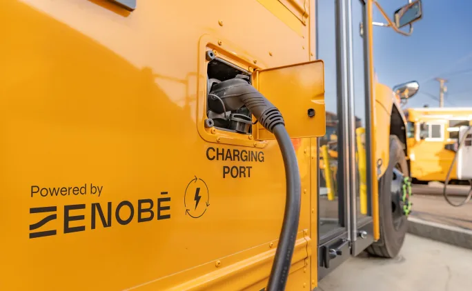 Close-up of a yellow electric school bus charging port with cable plugged in, labeled ‘Powered by Zenobē