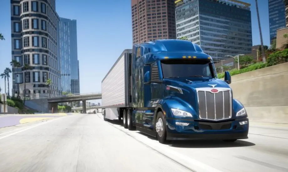 A blue Peterbilt Model 579 semi-truck driving on a city highway with tall buildings in the background.