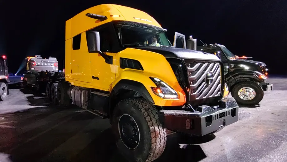 Yellow Volvo VNS heavy-haul sleeper truck with rugged tires and prominent grille, parked at night on a snow-covered lot beside a black truck, with a Volvo construction machine on a trailer behind it.