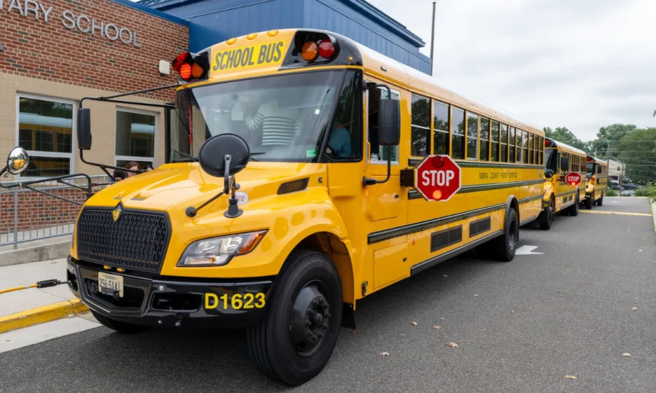 Yellow Fairfax County Public Schools buses line up outside an elementary school, with the lead bus displaying its extended stop arm and flashing red lights.
