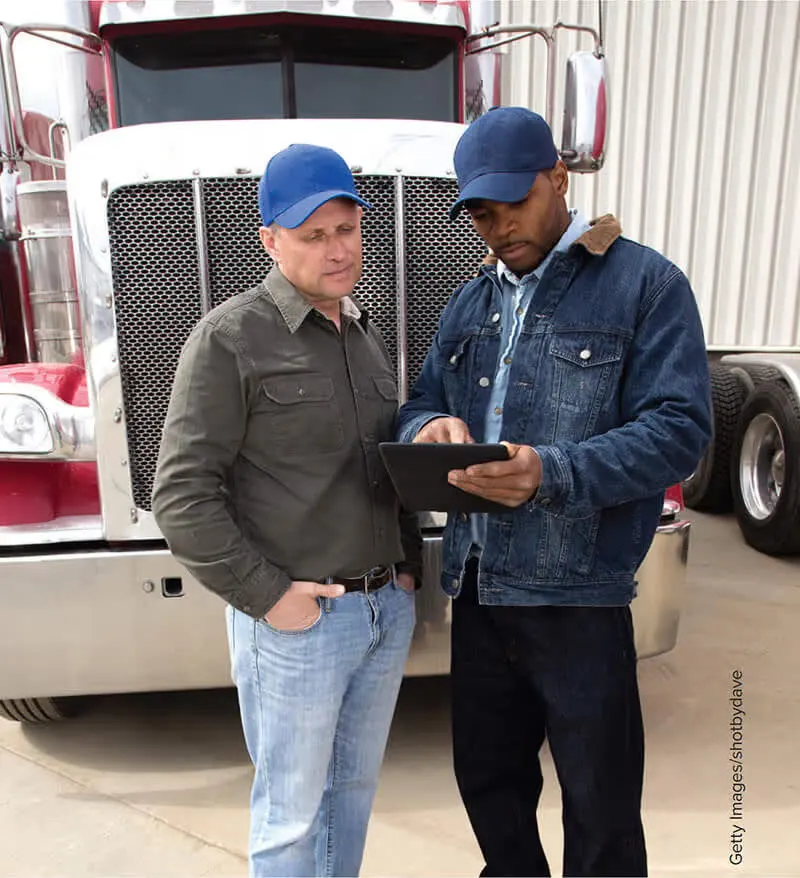 Two truck fleet managers standing in front of a semi-truck reviewing emergency response plans on a digital tablet.
