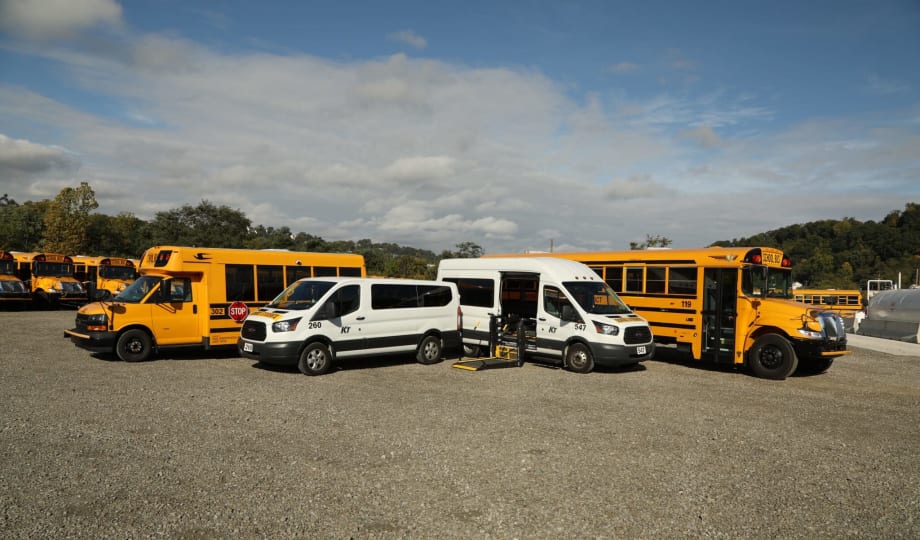 A lineup of Krise Transportation vehicles, including Type A and conventional school buses and passenger vans, with one wheelchair-accessible van shown with its lift extended.