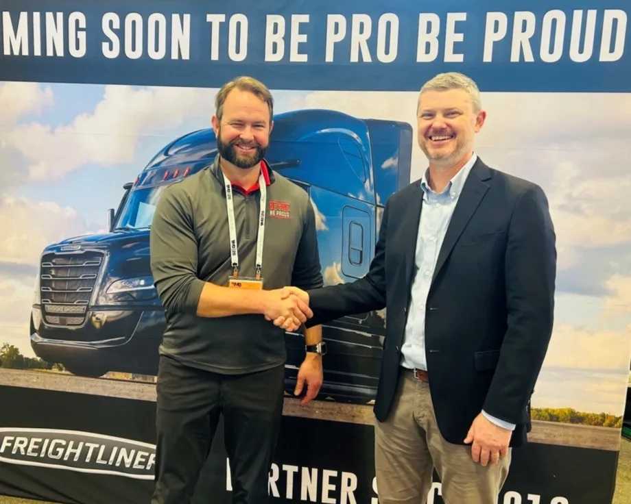 Two men shake hands in front of a Freightliner truck display during a partnership announcement supporting workforce development and training programs.