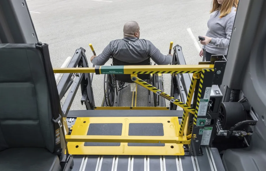 A wheelchair user operates a school bus wheelchair lift equipped with BraunAbility’s LiftSentinel barrier system as another person stands nearby with a handheld control.