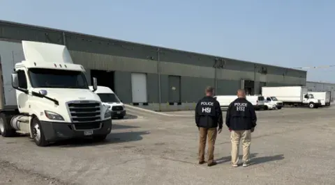 Two men in jackets with Homeland Security indicated on the back, standing in a parking lot looking toward a building with trucks in front of it