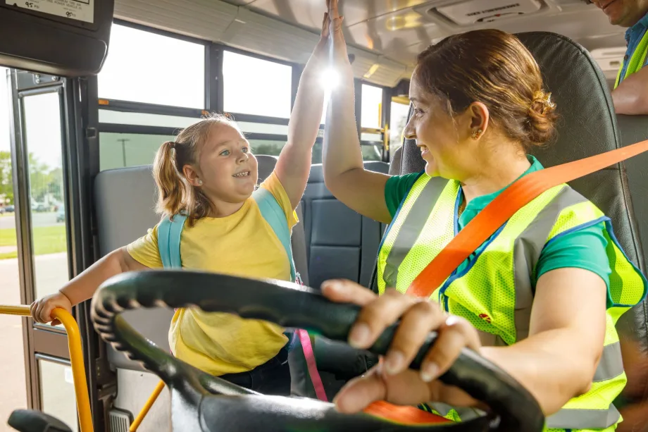 A school bus driver wearing a safety vest exchanges a high-five with a smiling young student near the front of a school bus. Sunlight streams through the windows as another adult stands nearby.