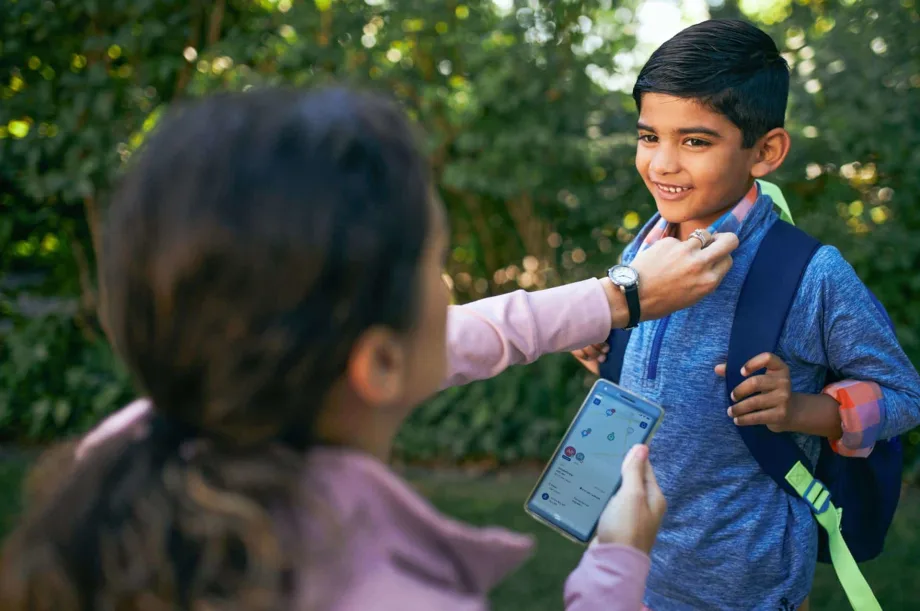 A parent uses the FirstView mobile app while helping a student with his backpack, illustrating real-time school transportation tracking and communication.