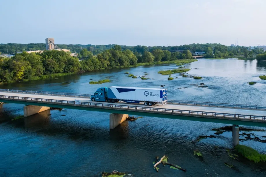A GP Transco tractor-trailer crosses a bridge over a river, with trailer side skirts visible beneath the white trailer.