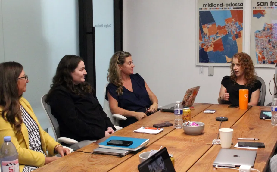 Four women participate in a council meeting around a wooden conference table, with one speaking while the others listen. Laptops, notebooks, and drinks sit on the table.
