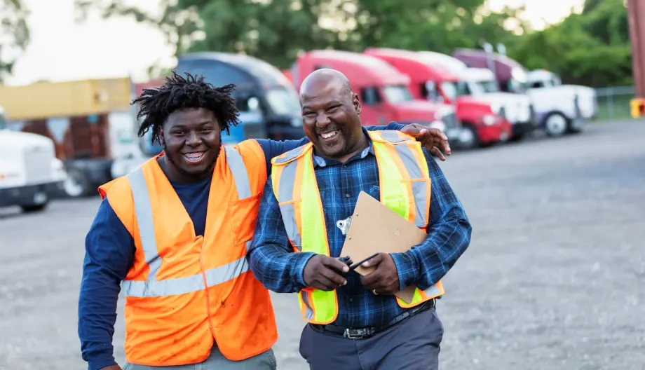 Two black men in safety vests walking together laughing in a truck fleet yard