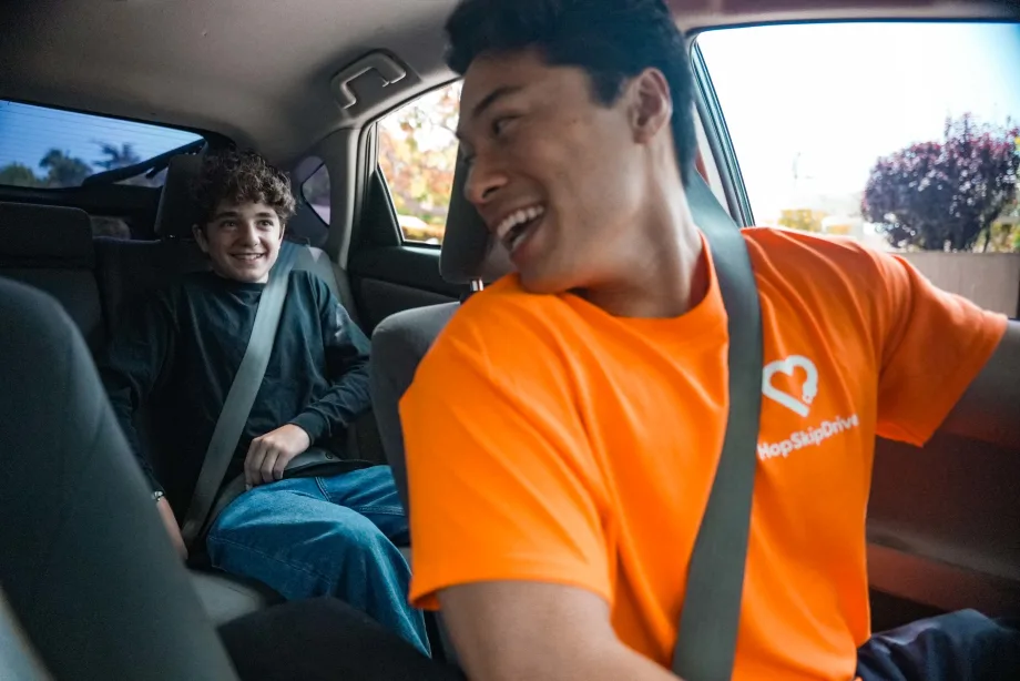 HopSkipDrive driver in an orange shirt smiles at a seat-belted student riding in the back seat of a car.