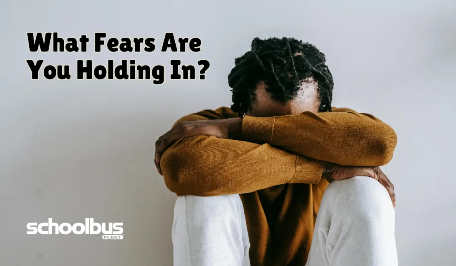 A person sits on the floor with their knees pulled in and arms crossed over them, head resting down on their arms, conveying stress or anxiety. Text on the image reads, “What Fears Are You Holding In?” with the School Bus Fleet logo in the corner.