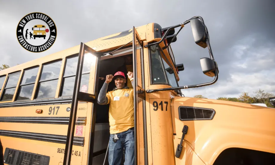 School bus driver stands in the doorway of a yellow school bus during the New York School Bus Contractors Association roadeo event, with dark storm clouds in the background and the NYSBCA logo in the corner.