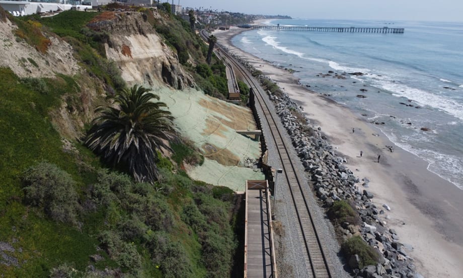 Coastal railroad tracks run between a steep bluff and the Pacific Ocean in San Clemente, California, with rock barriers protecting portions of the shoreline.