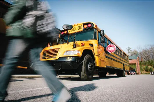 A yellow school bus with its red stop arm extended is stopped to load or unload students as a blurred child crosses in the foreground, illustrating the importance of motorists stopping for school buses and protecting students in the loading zone.