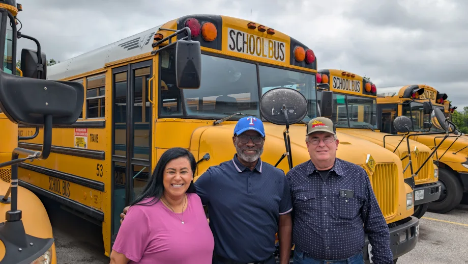 Three people pose in front of a line of school buses.