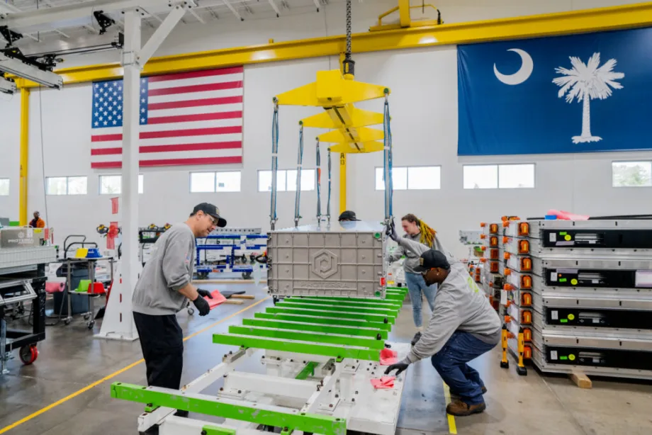 Workers assemble a large Proterra EV battery pack inside a manufacturing facility, using an overhead crane to position the battery module onto a chassis frame. American and South Carolina state flags hang above the production floor, with additional battery packs stacked nearby.
