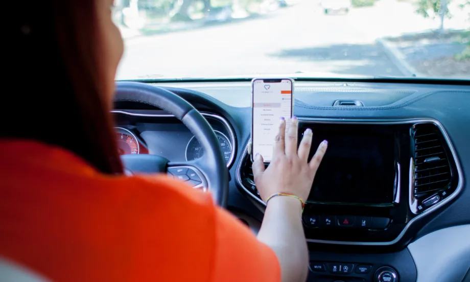 A driver in an orange t-shirt uses a smartphone mounted on a vehicle dashboard to access HopSkipDrive’s app.