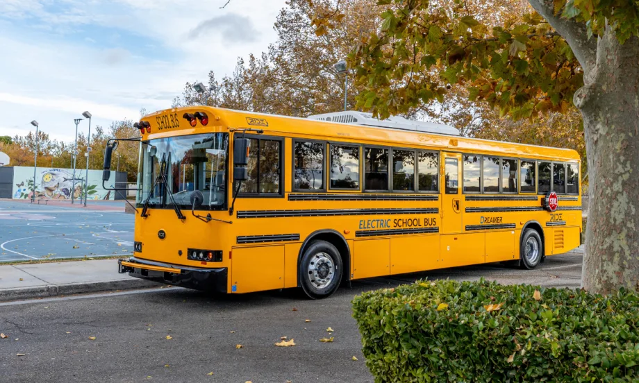 A yellow RIDE Type D electric school bus is parked along a tree-lined street outside a school.