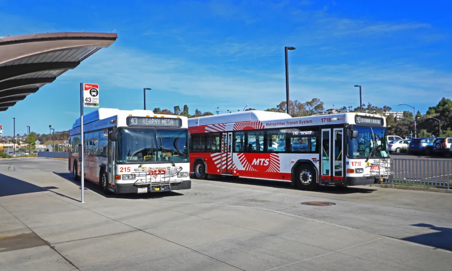 Two San Diego Metropolitan Transit System buses sit at a transit station under a clear blue sky.