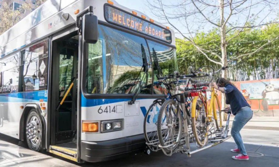 A passenger loads a bicycle onto the front bike rack of a VTA transit bus stopped along a city street.