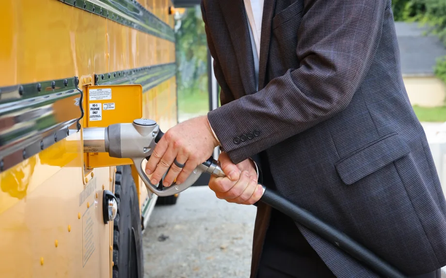 Close-up of a person refueling a yellow school bus with propane. The fuel nozzle is connected to the bus's propane fueling port, illustrating an alternative fuel option for school transportation fleets.