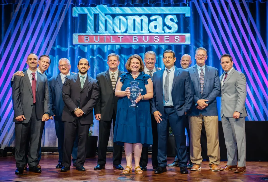 Group of Thomas Built Buses executives, employees, and award recipients pose onstage during an awards ceremony beneath a large Thomas Built Buses sign, with one woman holding a glass trophy at center stage.