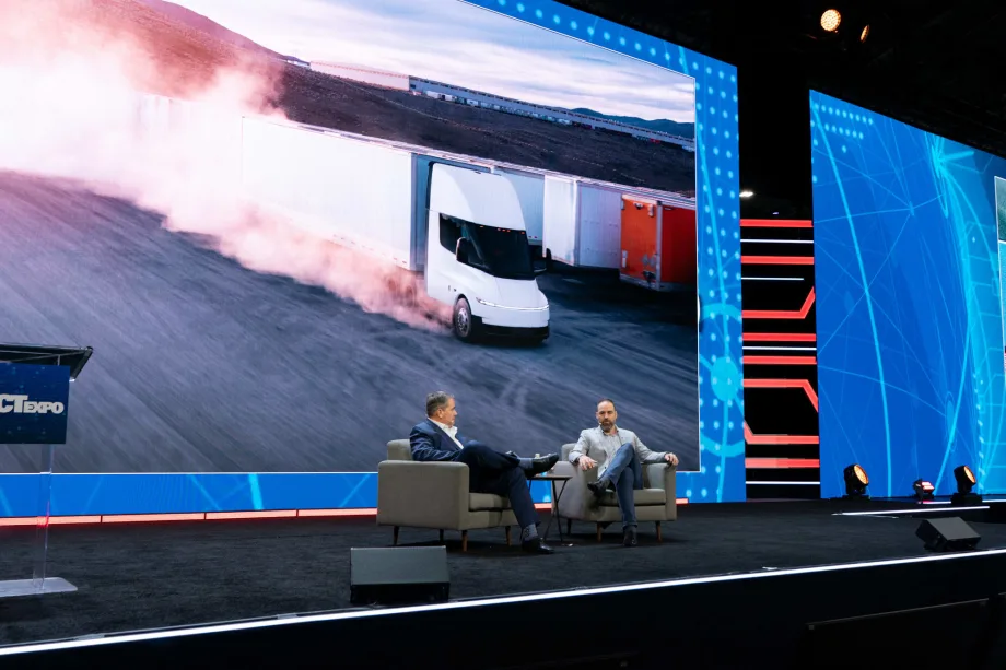 Two men in chairs on stage with big video screen behind them showing Tesla Semi