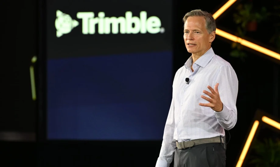 Photo of man in collared buttoned shirt speaking on stage in front of a backdrop that says Trimble